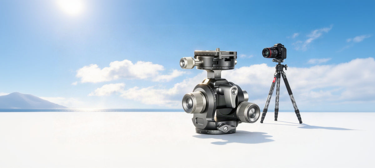 A black and silver camera tripod head shown prominently in the foreground, with a camera mounted on a separate tripod standing behind it against a bright blue sky with white clouds.